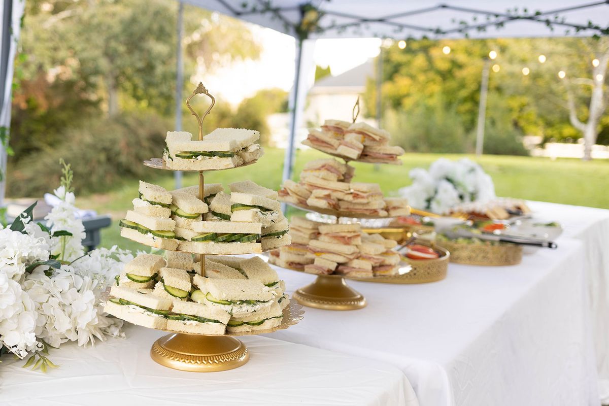 Refreshment table under a white canopy with tiered stands of tea sandwiches and pastries, with a lawn and trees behind