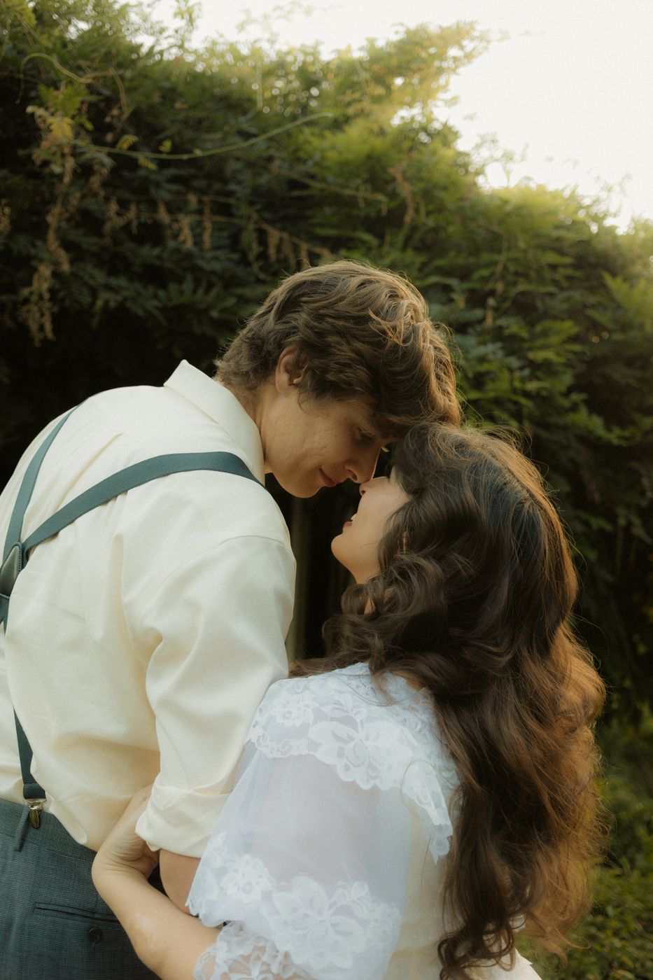 Close portrait of a couple touching foreheads in front of soft green foliage, the bride in white lace
