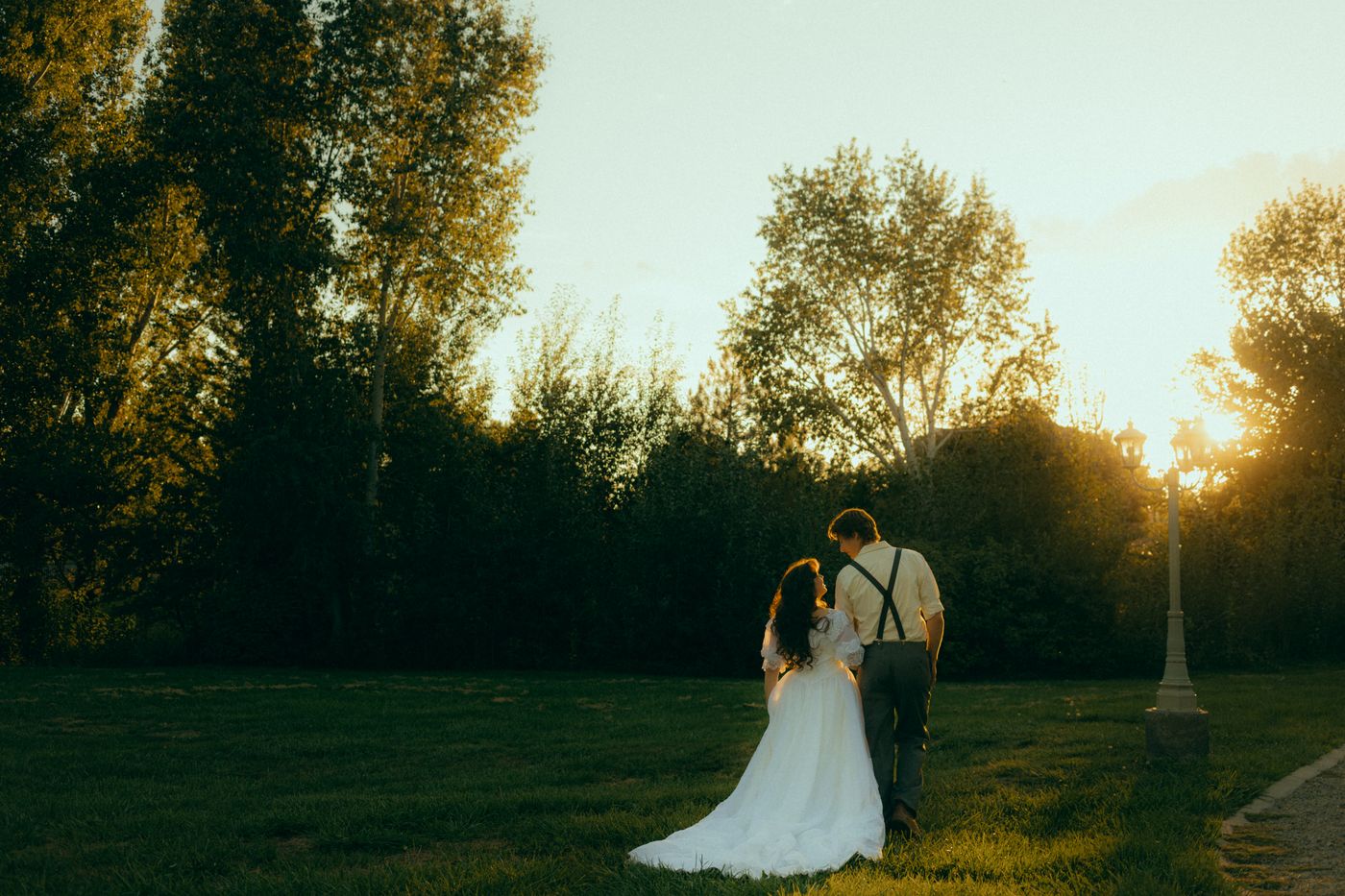 Couple standing together on the open lawn at golden hour, with sunlight glowing through tall trees and a lamppost