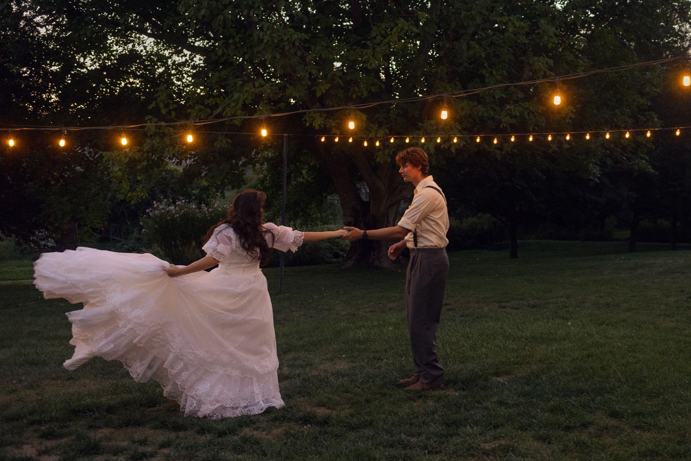 Bride in a flowing white gown twirling hand in hand with her partner on the lawn beneath glowing string lights at dusk