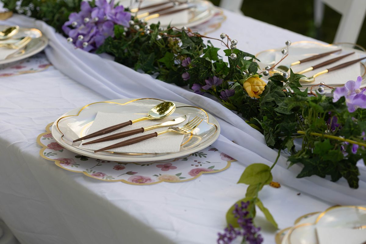 Table set with floral-rimmed plates, gold flatware, and a runner of greenery and purple flowers