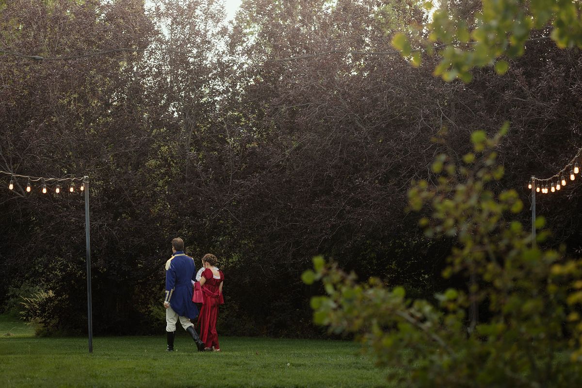 Couple in formal dress on the lawn at dusk between posts strung with lights, framed by dark trees