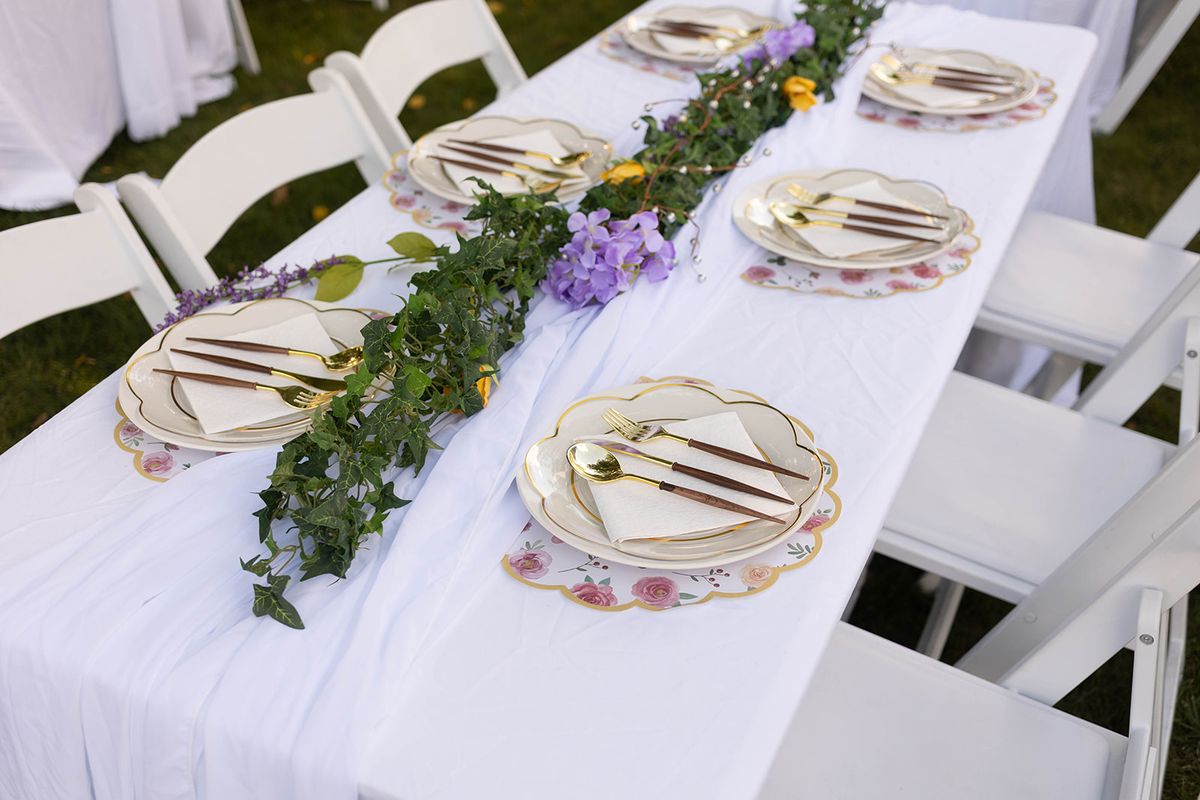 Long banquet table with white linens, floral-rimmed plates, gold flatware, and a greenery and purple flower runner, lined with white chairs